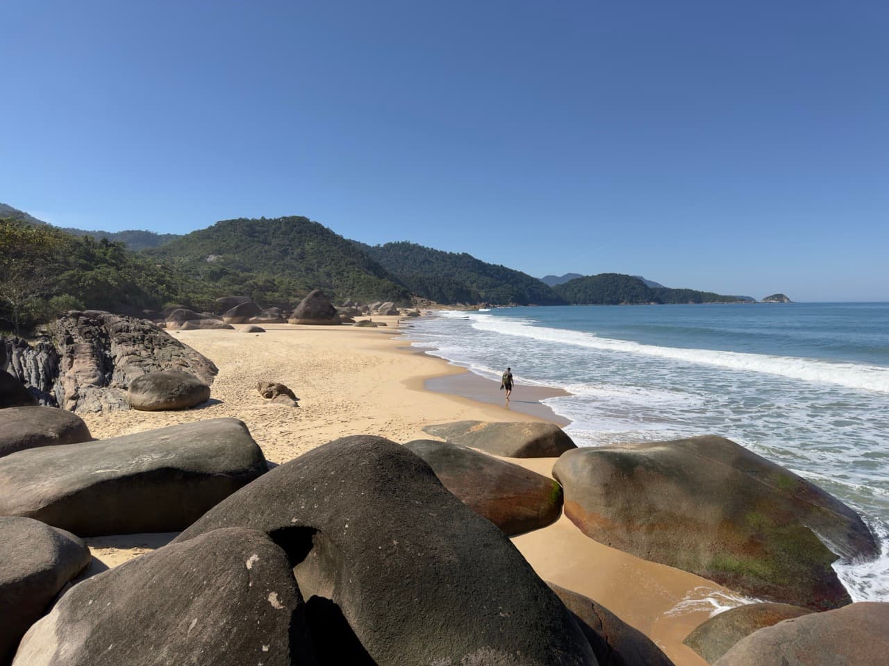 A person walking an empty beach framed by giant boulders and forested hills
